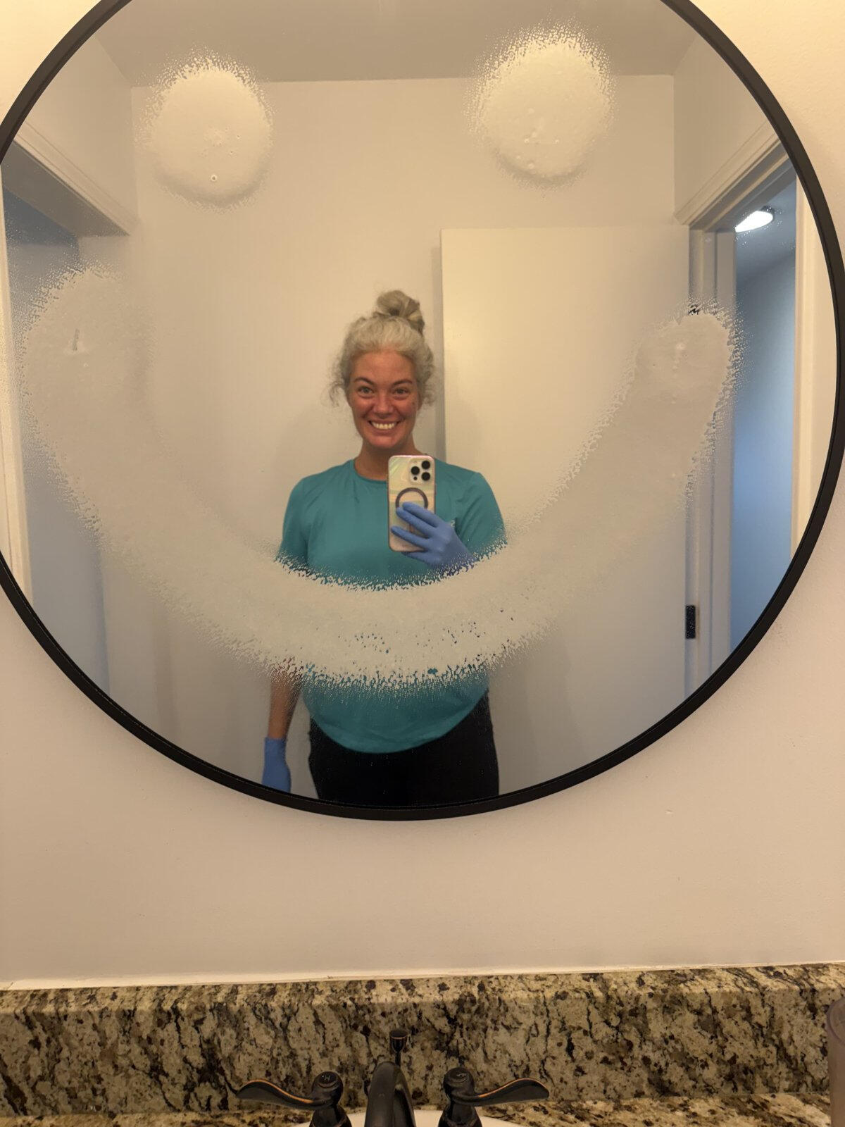 Shelbie Pickar smiling in a freshly cleaned bathroom mirror with a smiley face drawn in the cleaner